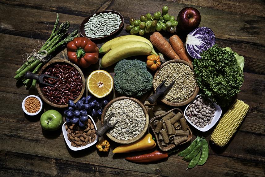 Table top view of fresh vegetables and legumes on rustic wooden table, including corn, avocado, broccoli, grapes, bell pepper, lettuce, banana, apple almonds and wholegrain pasta.