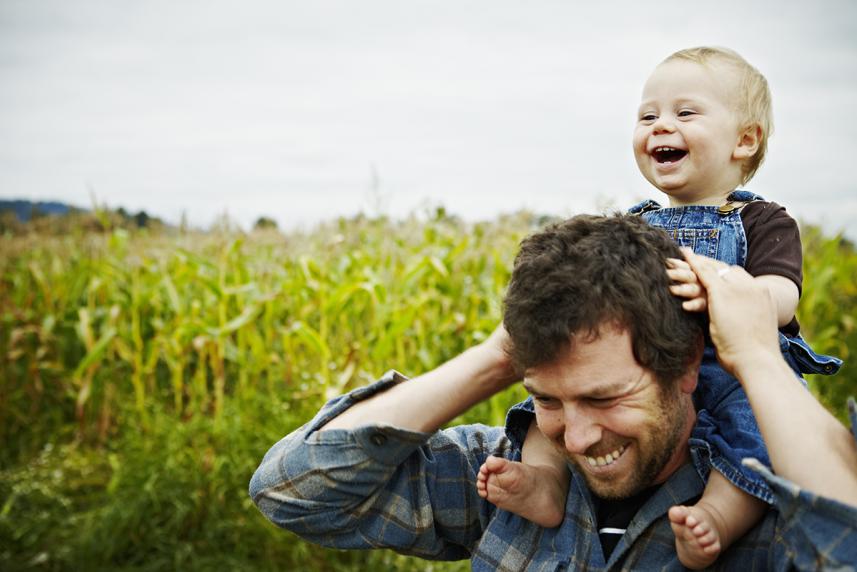 Farmer holding baby boy on shoulders laughing standing next to organic corn field