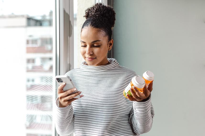 A young person standing by the window holding a smart phone and pill bottle researching medicine online