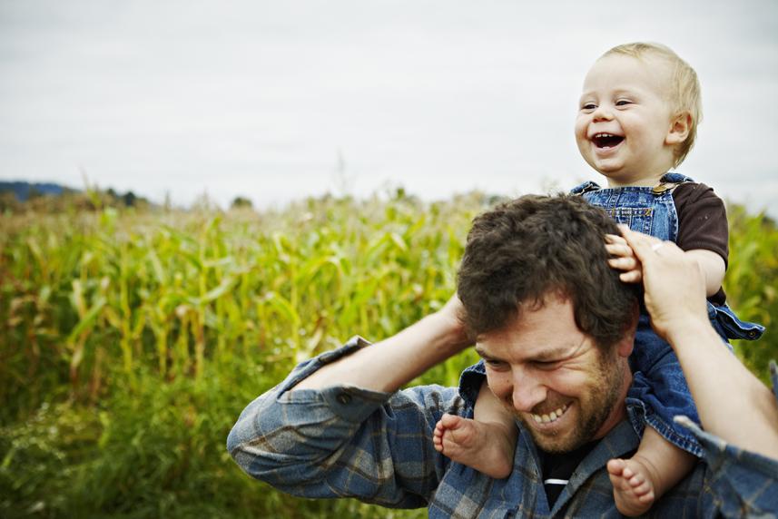 Farmer holding baby boy on shoulders laughing standing next to organic corn field