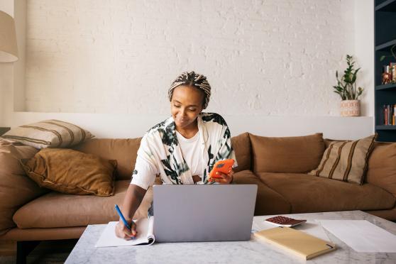 A person sits at a coffee table in a living room, looking at their phone while writing in a notebook beside an open laptop.