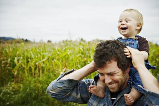 Farmer holding baby boy on shoulders laughing standing next to organic corn field